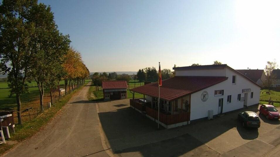 Blick auf ein weißes Gebäude mit rotem Dach, umgeben von Bäumen und einem Straßenverlauf in ländlicher Umgebung.