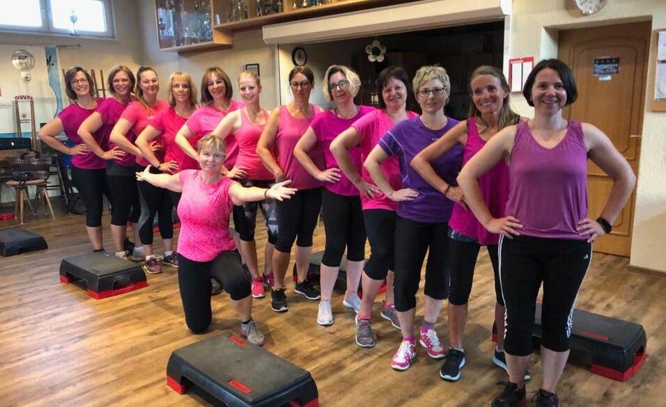 Gruppe von Frauen in Sportkleidung mit Rosatönen posiert lächelnd vor Step-Trainingsgeräten in einem Fitnessraum.